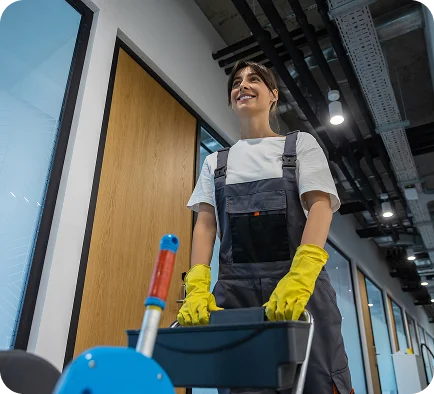 A smiling person in a jumpsuit and yellow gloves pushes a cleaning cart down a modern, well-lit office corridor, exuding professionalism and a positive attitude.