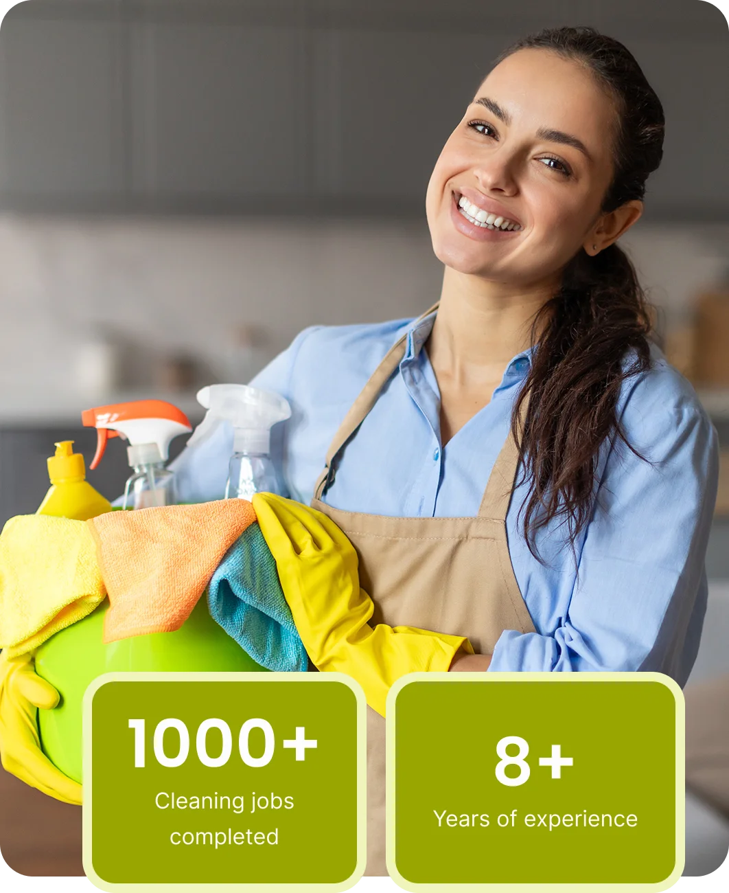 A smiling woman in an apron and yellow gloves holds a green bucket with cleaning supplies in a cozy kitchen. This scene conveys cleanliness and a positive attitude.