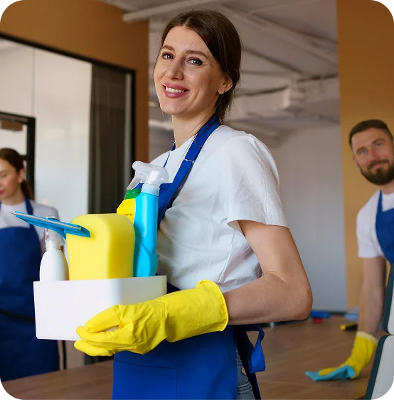 A smiling woman in a blue apron holds cleaning supplies along with two colleagues in yellow gloves in a bright office, highlighting teamwork.
