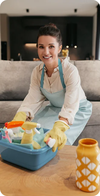 A smiling woman in a blue apron and yellow gloves holds a basket of cleaning supplies. She is in a cozy living room that radiates a joyful atmosphere and order.